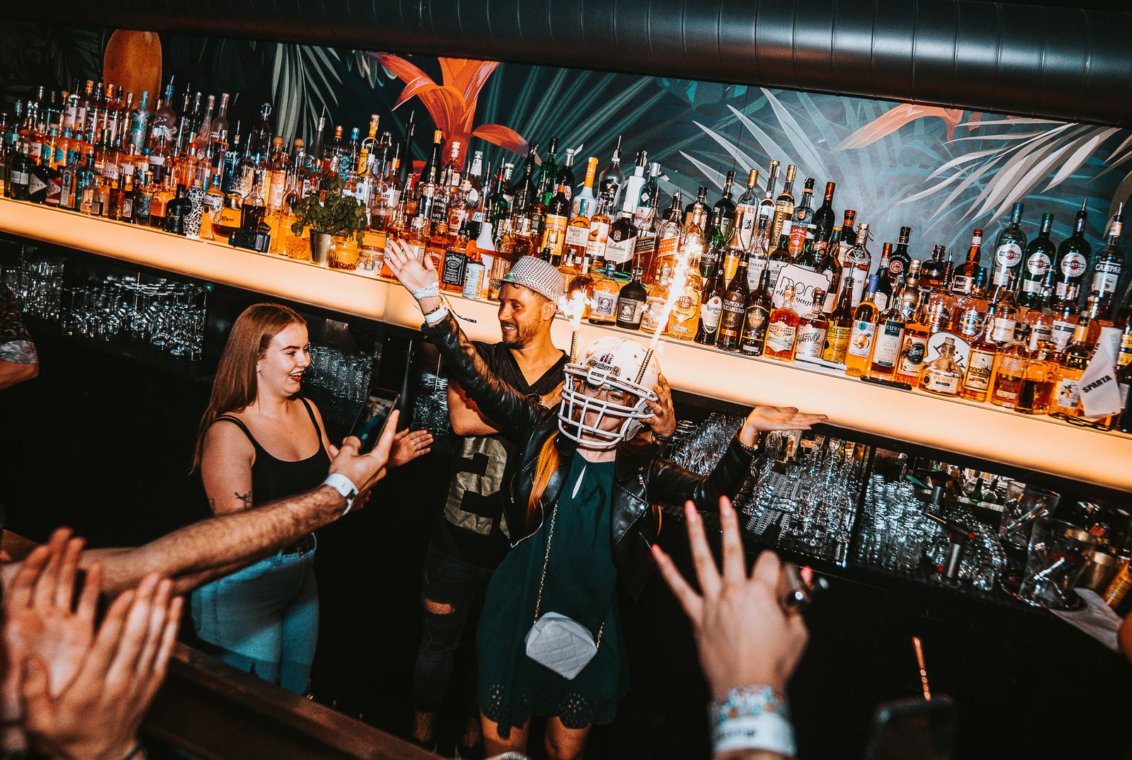 Bar Mango: nasvícená stěna s lahvemi, barman a hosté u baru